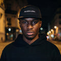 Person wearing a black hoodie and cap with 'MAGDALENA' text, standing on a street at night.