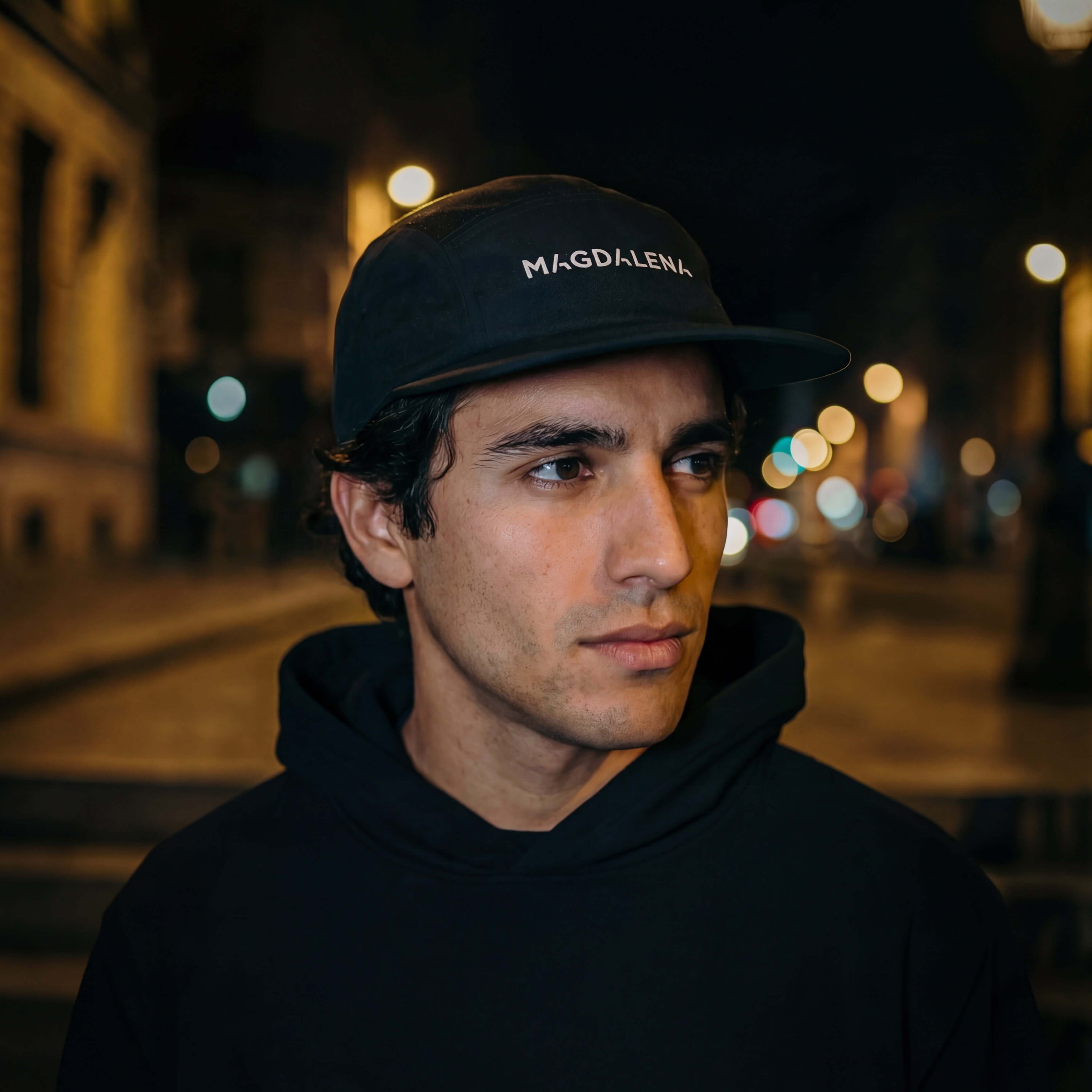 Man wearing a black cap with 'Magdalena' text in an urban night setting