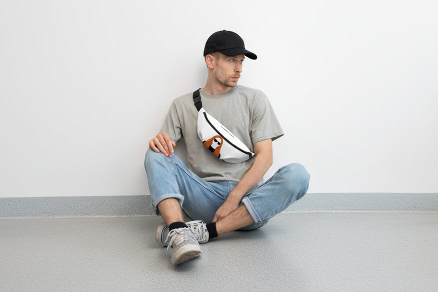Man sitting on a step wearing a gray t-shirt, black cap, and blue jeans with a white fanny pack featuring a logo.