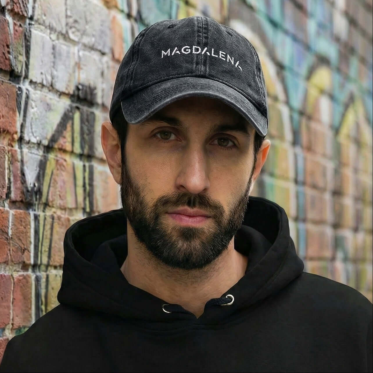 Man wearing a black hoodie and cap with 'Magdalena' branding against a graffiti-covered wall.