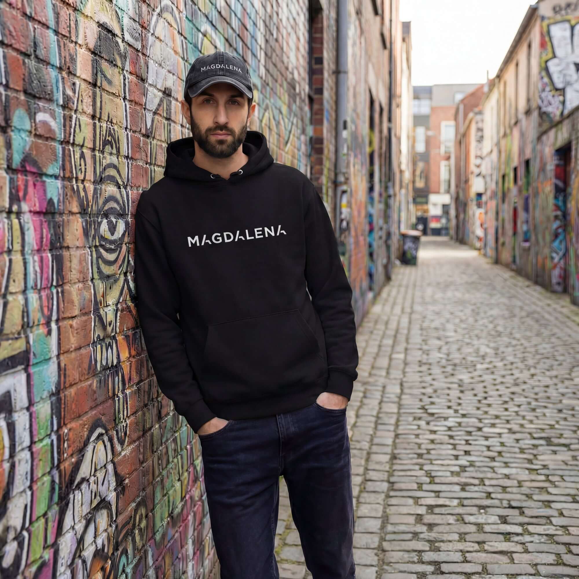 Man wearing a black hoodie with 'MAGDALENA' text in a graffiti-covered alley.