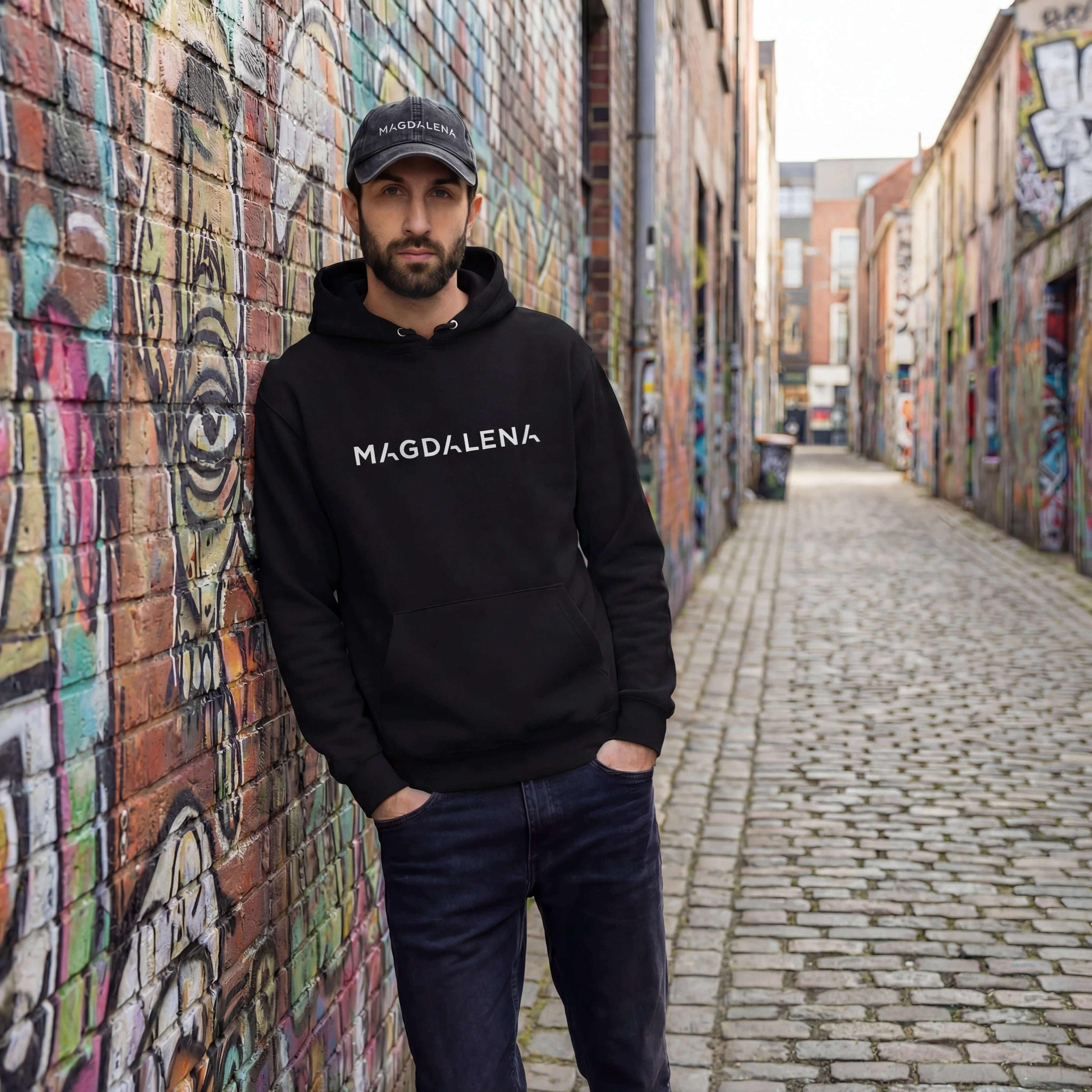 Man wearing a black hoodie with 'MAGDALENA' text in a graffiti-covered alley.