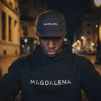 Person wearing a black hoodie and cap with 'MAGDALENA' text, standing on a city street at night.