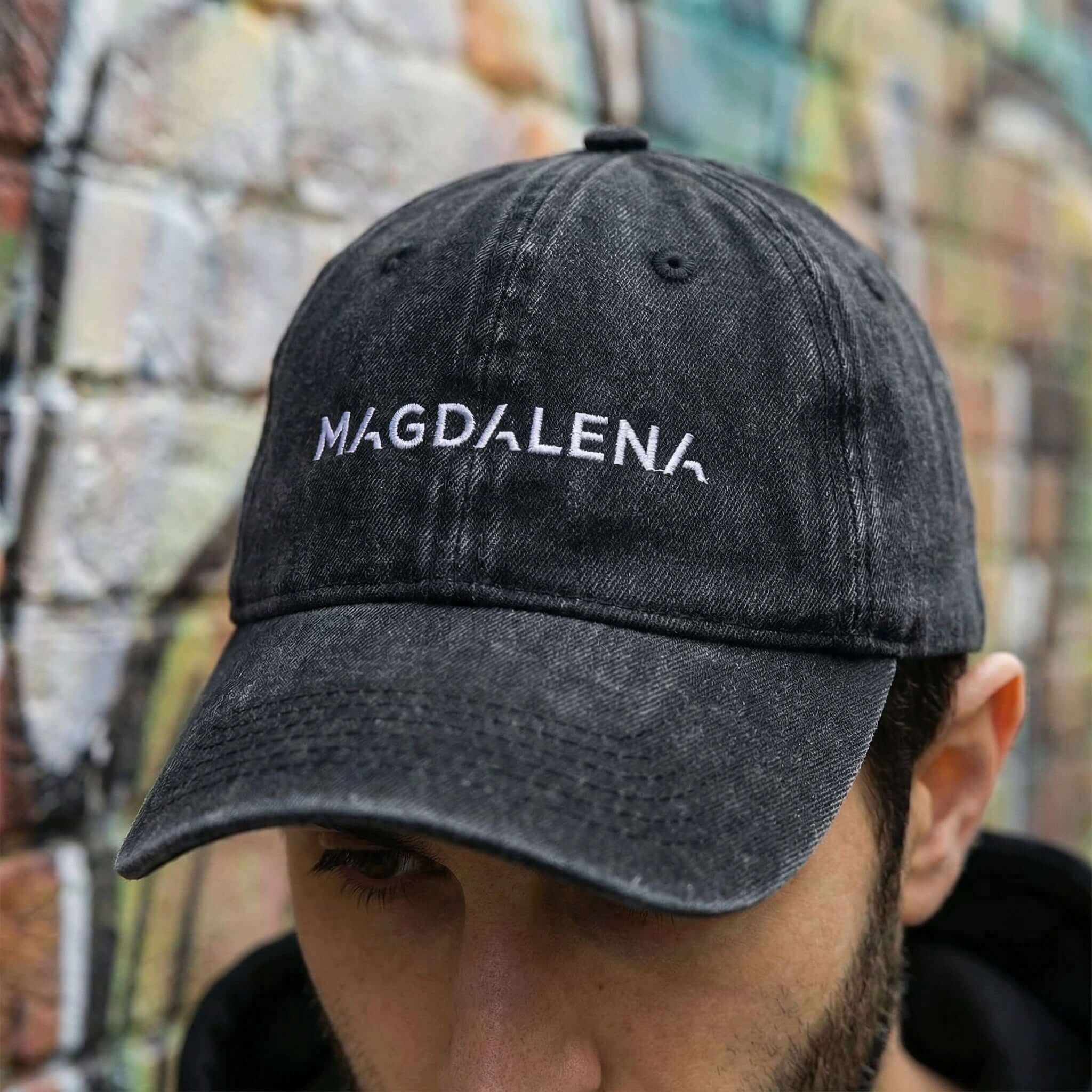 Black cap with 'MAGDALENA' text worn by a person against a graffiti-covered wall.